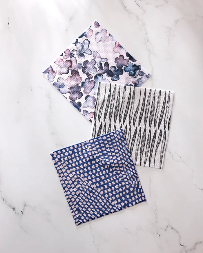 Three GEOMETRY Tiny Petals dishcloths—featuring purple and blue florals, black wavy lines on white, and a blue-white grid—are made from super-absorbent recycled materials and displayed on a white marble surface.