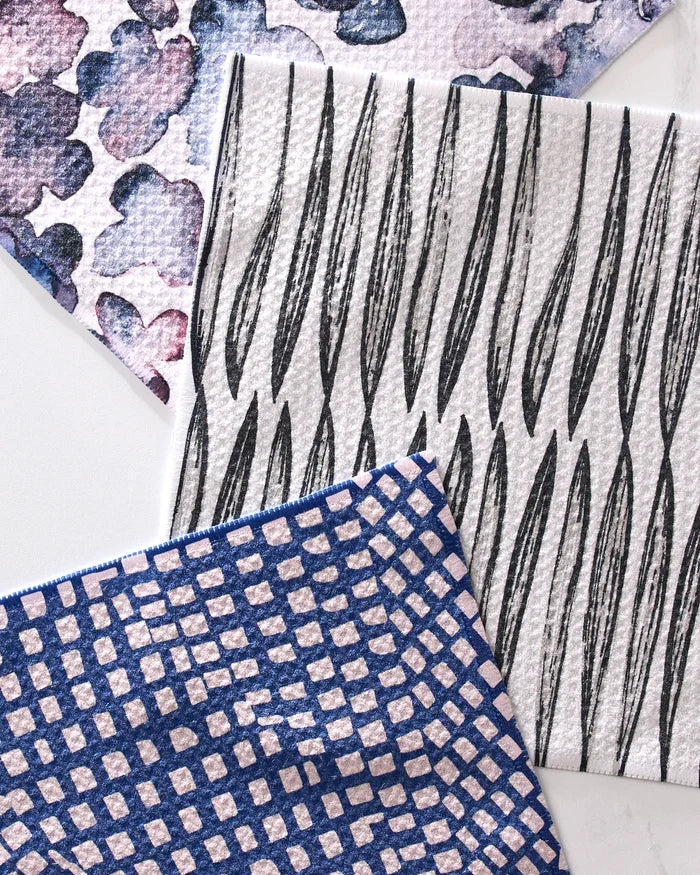 Three pieces from GEOMETRY’s Tiny Petals set—a purple watercolor-spotted, a black leaf-patterned, and a blue-and-white geometric towel—overlap on a white surface. These super-absorbent towels are made from recycled materials.