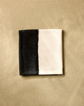 A neatly folded square napkin, inspired by the GEOMETRY Top Layer, features one half black and the other half white, resting on a sandy beige surface.
