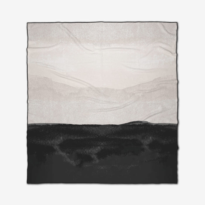 The GEOMETRY Top Layer is a square recycled beach blanket with a minimalist landscape design, featuring a black land-like foreground and light gray and white mountains and sky. Visible fabric creases add an organic touch.