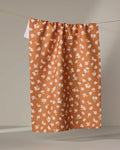 The Tossed Wildflowers Rust tea towel by Mandy Arroyo features a rust-orange color, a scattered wildflower print in white, pink, and yellow, and is made from recycled materials, shown hanging vertically against a neutral background.
