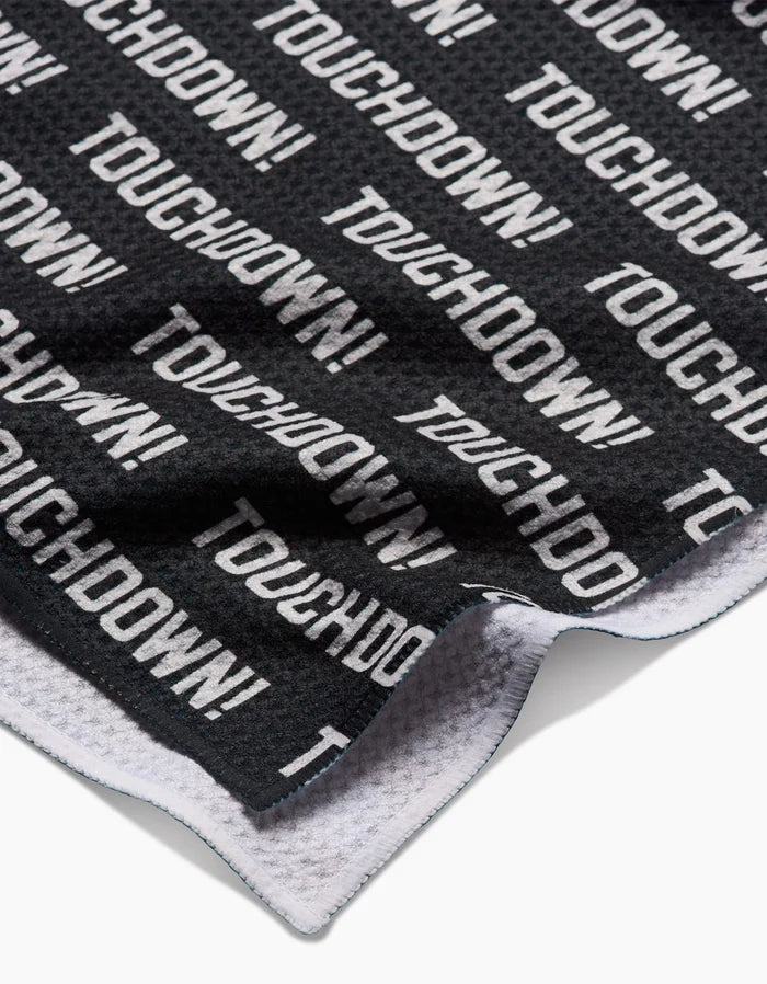 A close-up of a black GEOMETRY Geoweave Touchdown Tea Towel shows TOUCHDOWN! in bold white letters. The edge is slightly folded to reveal the white underside, and its made from recycled materials.