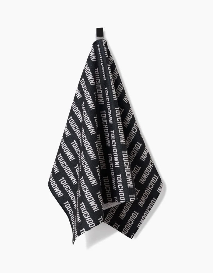 The GEOMETRY Touchdown tea towel, crafted from Geoweave fabric in black with TOUCHDOWN repeated diagonally in white, hangs by a black loop against a plain white background.