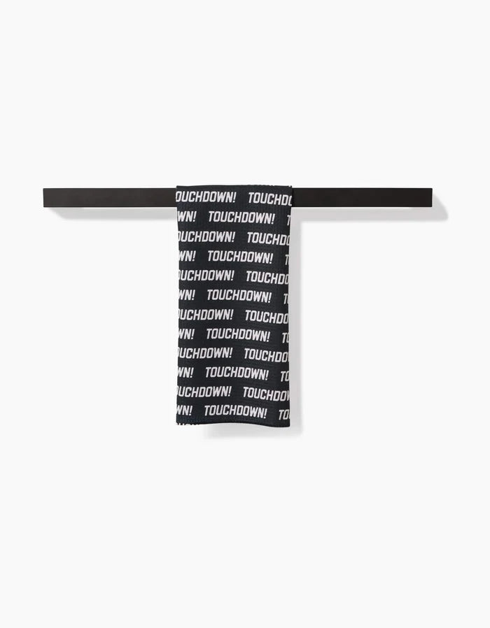 The GEOMETRY Touchdown tea towel, made from recycled materials, features bold black and white TOUCHDOWN! text and hangs neatly on a horizontal black rack against a white background.