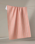 The Twig + Spark "Towering Twigs" towel, made from recycled materials, features a subtle white branch pattern on pink and hangs from a string against a neutral background.