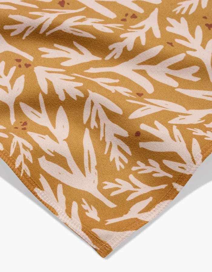 A close-up of Twig + Sparks Trail Dusting Gold napkin, a brown absorbent cloth with a white abstract leafy pattern and neatly stitched edge.