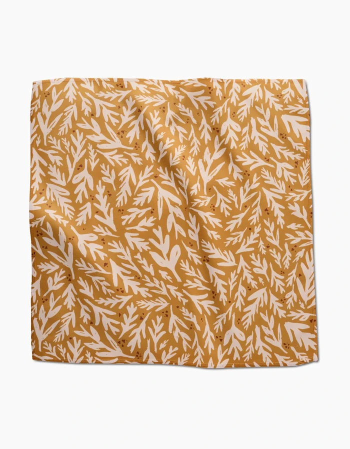 The Trail Dusting Gold napkin by Twig + Spark is a square, recycled cloth with a mustard yellow background and white leafy branch pattern. The absorbent napkin has a slightly wrinkled texture and is shown on a white surface.
