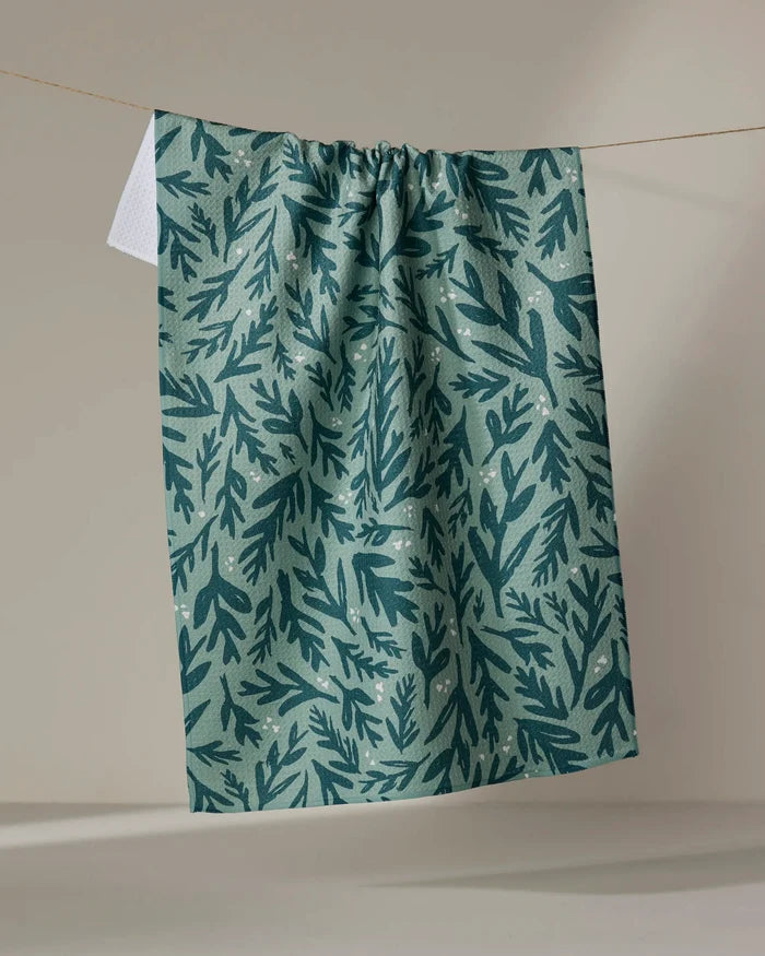 The Twig + Spark "Trail Dusting" green tea towel, featuring a dark green leafy pattern, hangs on a clothesline against a neutral background.