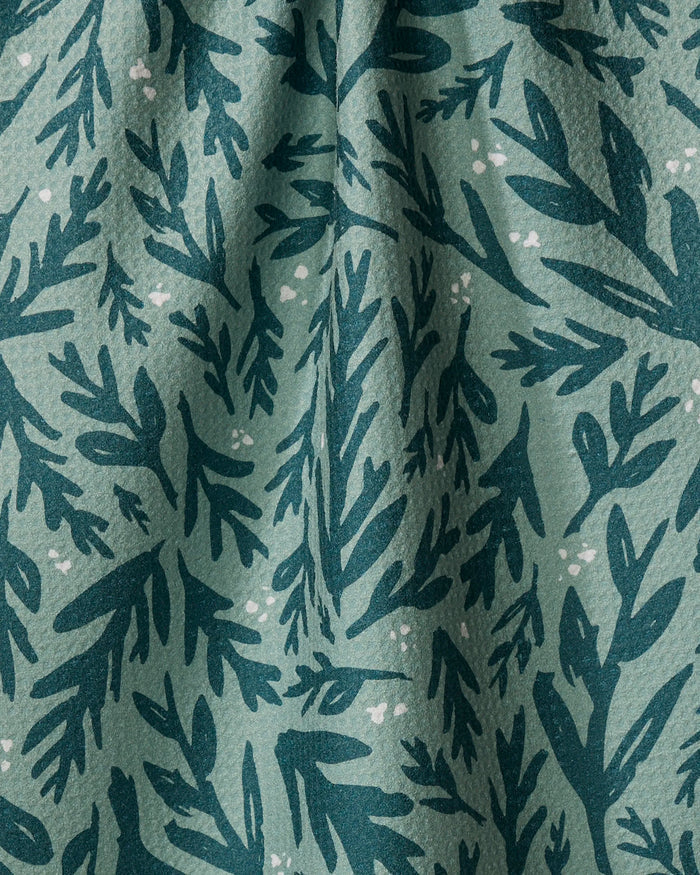 A close-up of Twig + Spark’s Trail Dusting recycled kitchen towel shows green fabric with a dark green leafy pattern, white dots, soft folds, and a textured surface.