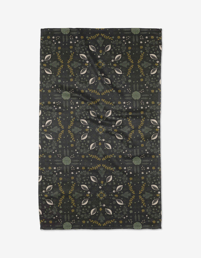 The Ash Cascade Trailhead tea towel is rectangular and dark, with a symmetrical leaf and botanical pattern in muted green, yellow, and beige. Made from eco-friendly fabric, it has gentle wrinkles and is shown flat on a white background.