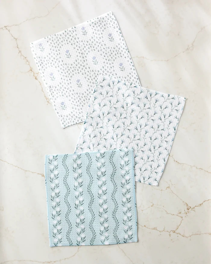 Three "Trailing Farmhouse Florals" napkins by Teri the Butter, featuring white and light blue botanical patterns and made from recycled materials, are arranged on a light marble surface.