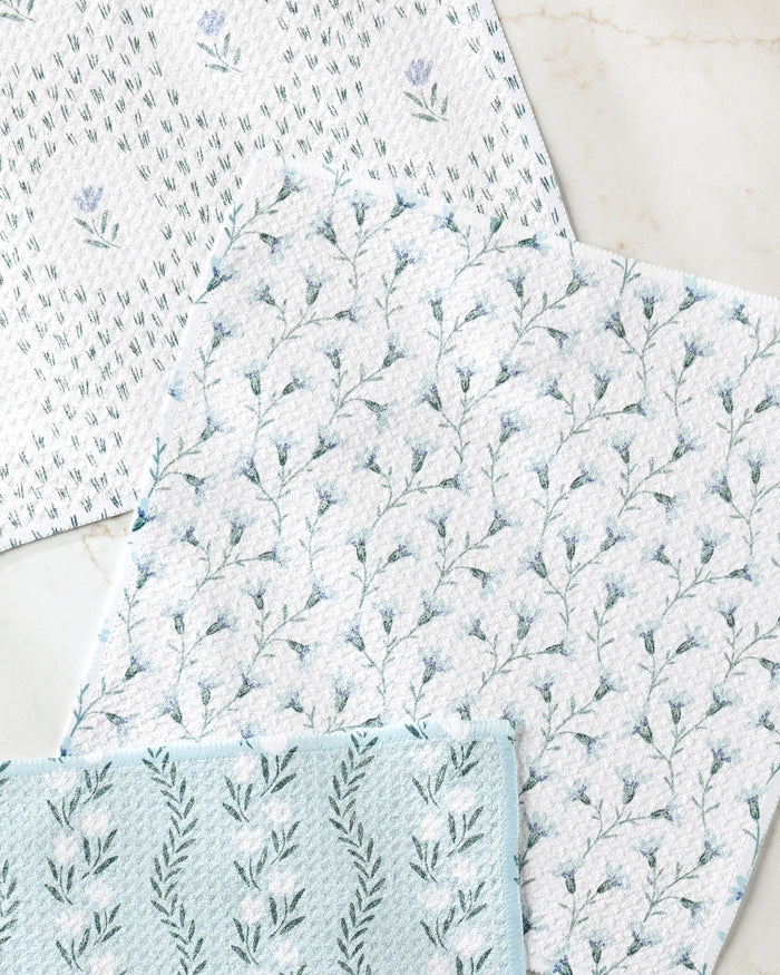 The Teri the Butter Trailing Farmhouse Florals set includes three absorbent dishtowels, each with a unique blue and green floral pattern featuring small flowers and leafy branches, displayed on a light marble surface.
