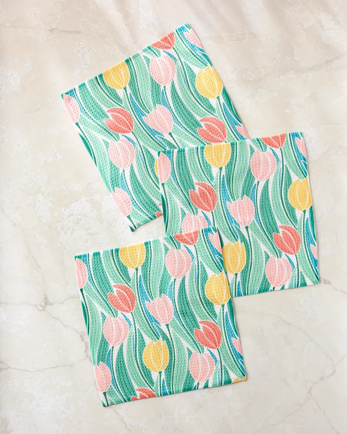 Three square Tranquil Tulips dishtowels by Sarah Daugherty, featuring a vibrant tulip design in yellow, pink, and red on a green and white background, displayed on light beige marble.