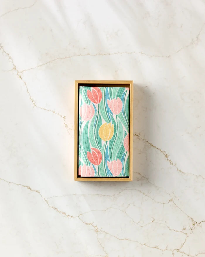 The Tranquil Tulips rectangular box by Sarah Daugherty, with a floral lid of pink and yellow tulips and green leaves, rests on a light marble surface and is designed to hold eco-friendly paper towels.