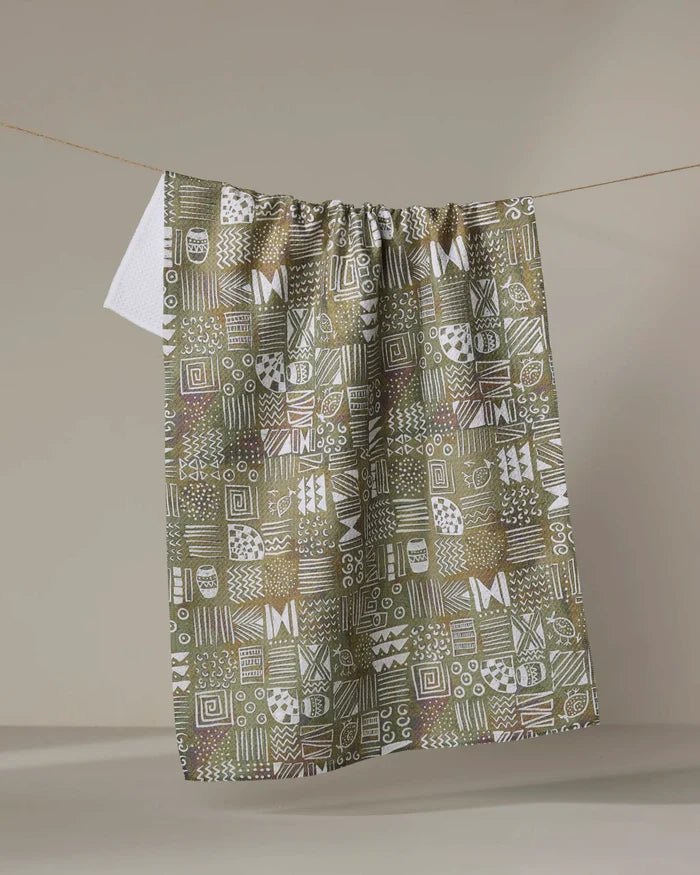 The "Tribal Symbols" tea towel by Taryn Brennan features a geometric abstract pattern in green, brown, and white. Made from recycled materials, it displays unique squares with lines, dots, and shapes against a beige background.