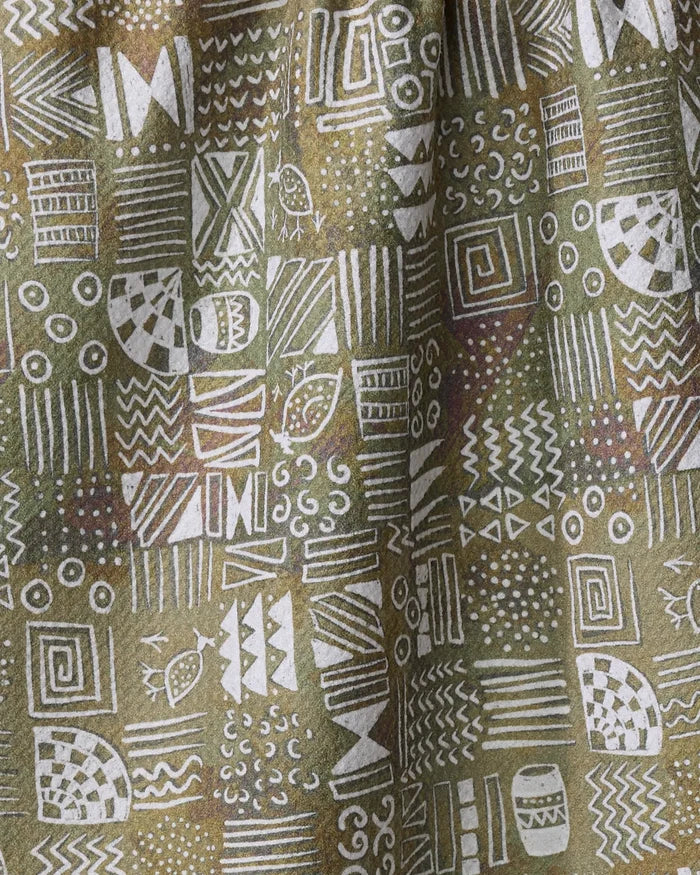 The Taryn Brennan "Tribal Symbols" kitchen towel features white geometric shapes, zigzags, spirals, dots, and lines in a grid over green and brown fabric with a slightly draped appearance.