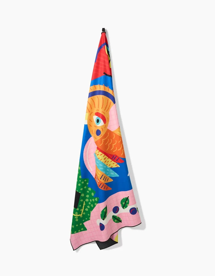 The Tropical Bird towel by Camila Leão hangs on a hook, quick-drying and colorful, showcasing a vibrant abstract design with an orange bird, green leaves, and bold pink and blue shapes.