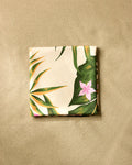 The GEOMETRY Tropical Jungle beach blanket, made from recycled materials, dries quickly and displays a tropical leaf and pink flower pattern on smooth light brown sand.