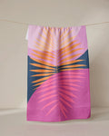 The GEOMETRY Tropical Summer tea towel, made with Geoweave fabric and featuring vibrant palm leaves on a purple and dark blue background, hangs on a clothesline against a neutral wall.
