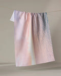 The Kait Hanson "Tropical Sunrise" towel, featuring a pastel gradient of pink, lavender, and blue, hangs textured and absorbent over a clothesline against a neutral backdrop.