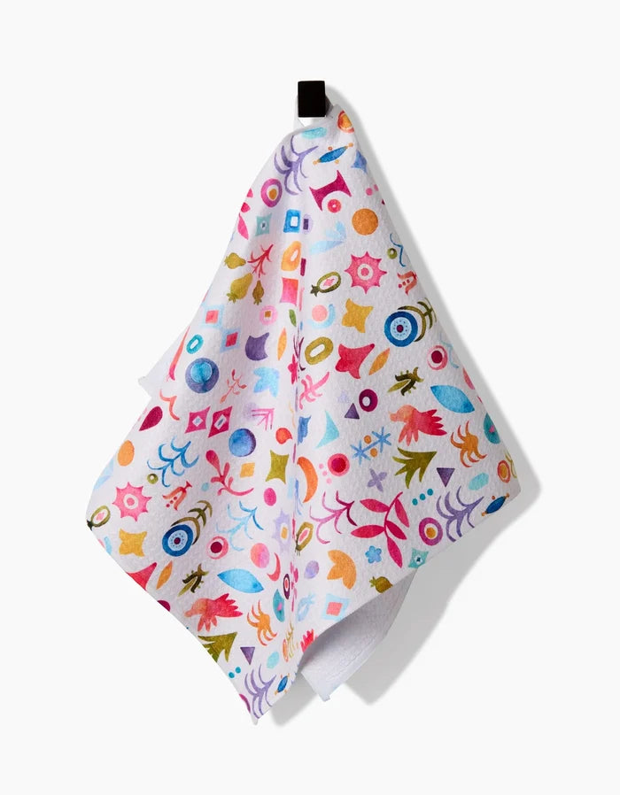 The Tula bar towel by Maayan Alper-Swan is white, made from recycled materials, and showcases colorful abstract shapes—eyes, leaves, and geometric patterns—with a black hanging loop against a plain background.