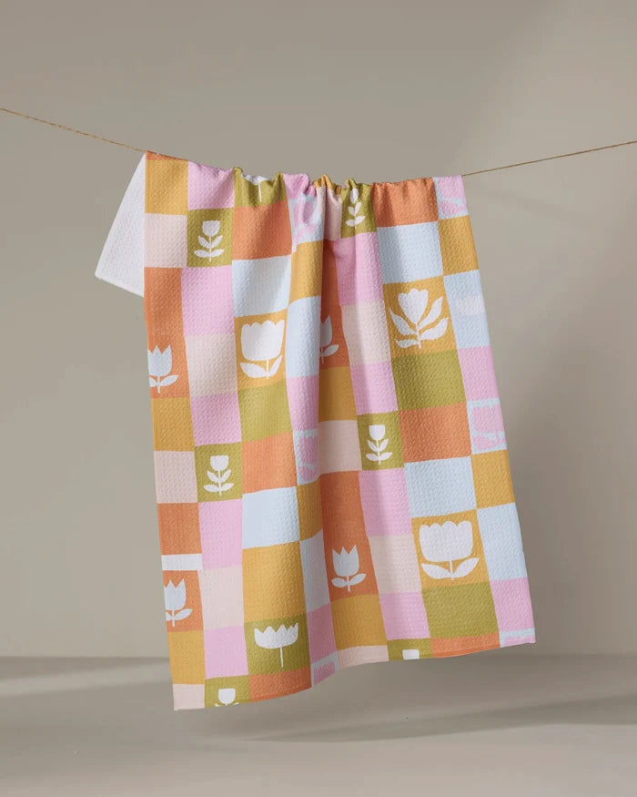 The Tulip Picnic tea towel by Lisa Nelson features pastel pink, yellow, orange, and green checkered patterns with white tulip and leaf motifs, shown hanging on a clothesline against a neutral background.