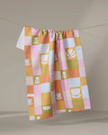 The Tulip Picnic tea towel by Lisa Nelson features pastel pink, yellow, orange, and green checkered patterns with white tulip and leaf motifs, shown hanging on a clothesline against a neutral background.