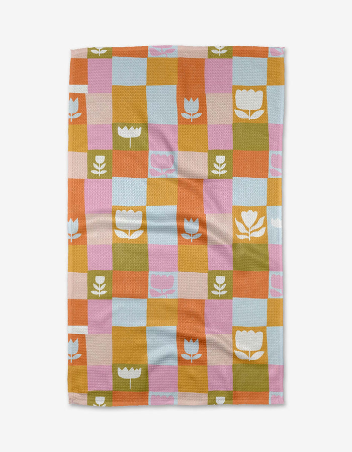The Tulip Picnic tea towel by Lisa Nelson features a checkered pattern in pastel blue, pink, orange, yellow, and green with white or pink floral and tulip designs. Made from recycled materials, it is shown laid flat on a white background.