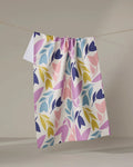The Lizzie Clark Tulip Tumble is a white, absorbent kitchen towel featuring a colorful leaf pattern in purple, pink, yellow, and blue. It’s pictured hanging on a clothesline against a plain, light background.