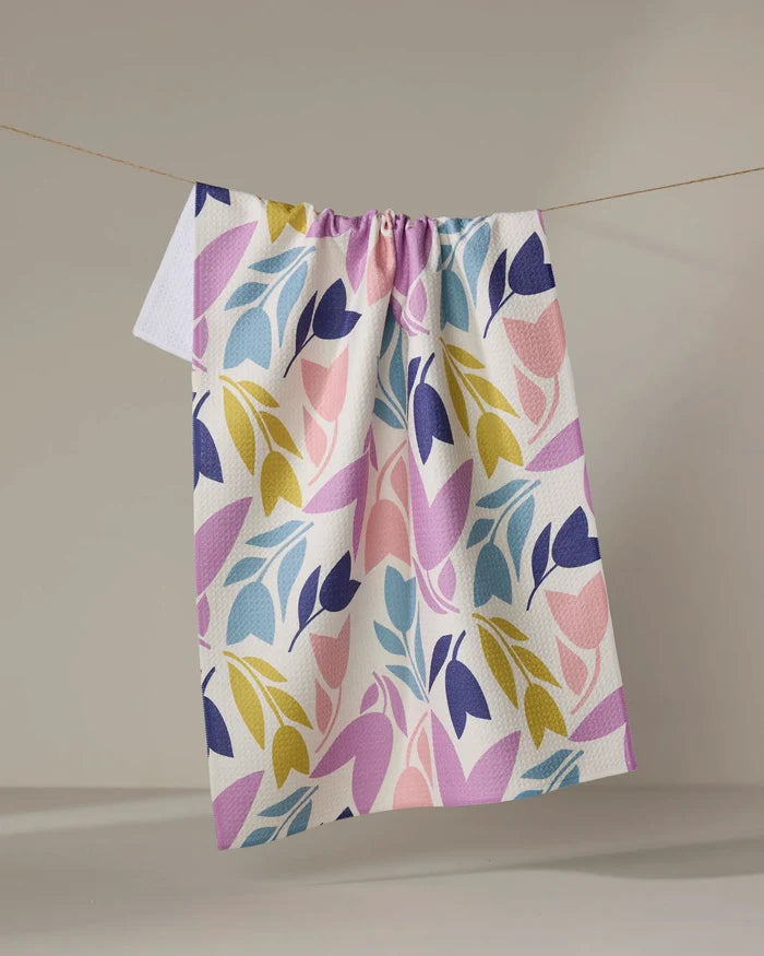 The Lizzie Clark Tulip Tumble is a white, absorbent kitchen towel featuring a colorful leaf pattern in purple, pink, yellow, and blue. It’s pictured hanging on a clothesline against a plain, light background.