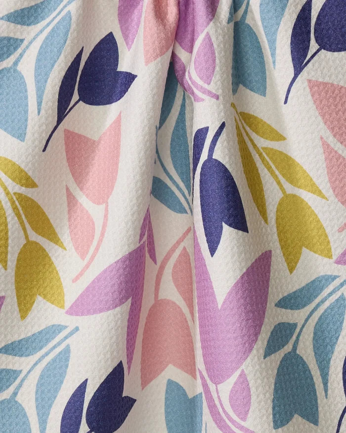 Bright and absorbent, the Tulip Tumble kitchen towel by Lizzie Clark features blue, yellow, pink, and purple floral patterns on a textured white background with a central fold.
