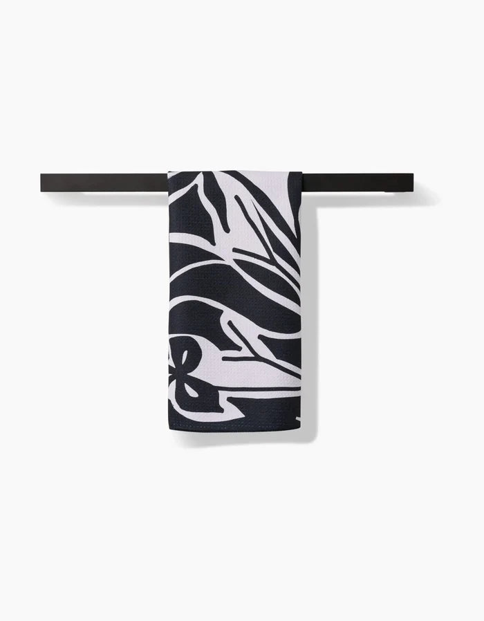 The Ash Cascade Twilight Plant kitchen towel, featuring a bold black and white abstract floral pattern, hangs neatly folded over a sleek black bar against a white background.