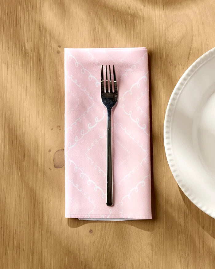 A silver fork rests on Elena Fay's Twirls pink recycled dinner napkin with a white pattern, atop a wooden table beside a scalloped-edge white plate.