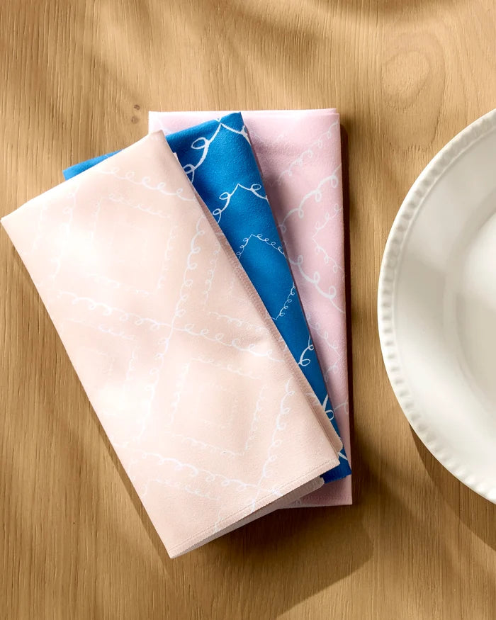 Three folded Twirls napkins by Elena Fay—two light pink and one blue with white geometric patterns—are arranged on a wooden table beside a white ceramic plate. Sunlight highlights these eco-friendly, recycled dinner napkins.