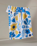 The Ukrainian Florals tea towel by Marina Solodka, featuring vibrant blue and yellow blooms and made from recycled materials, hangs on a clothesline against a neutral background.
