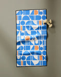 The Happy Hemera Ultra Marine Harmony towel, featuring a blue, white, and orange geometric pattern with abstract shapes and a wrinkled texture, hangs on a beige figure-shaped wall hook.