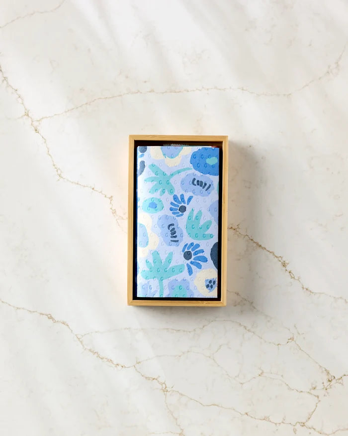 A small box from Sareka Unique sits on a light marble surface, containing "Unique"—an eco-friendly kitchen towel with abstract blue floral and leaf patterns in folded form.