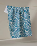 The Untangled tea towel by Rebecca Woolbright features a blue and white lotus-inspired pattern, crafted from recycled materials, and hangs on a clothesline against a plain beige background.