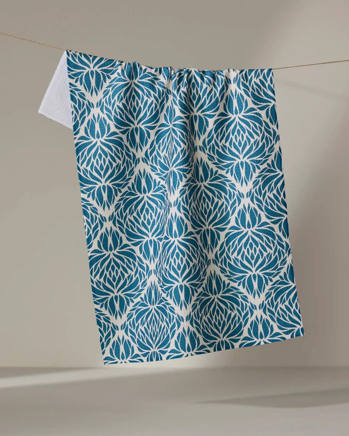 The Untangled tea towel by Rebecca Woolbright features a blue and white lotus-inspired pattern, crafted from recycled materials, and hangs on a clothesline against a plain beige background.