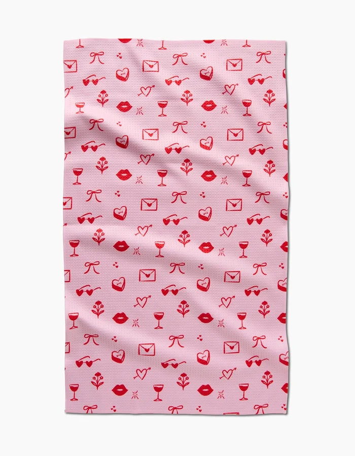 The V Day Doodles tea towel by Laura Supnik is made from recycled materials and features a playful, romantic design with red and pink icons like lips, hearts, envelopes, bows, leaves, wine glasses, and flowers.