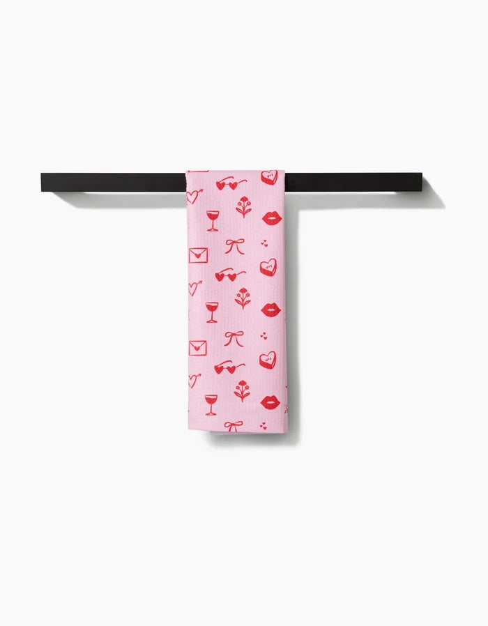 The V Day Doodles tea towel by Laura Supnik, made from recycled materials, displays red lips, hearts, bows, envelopes, and wine glasses on pink fabric. It hangs on a black towel bar against a white background.