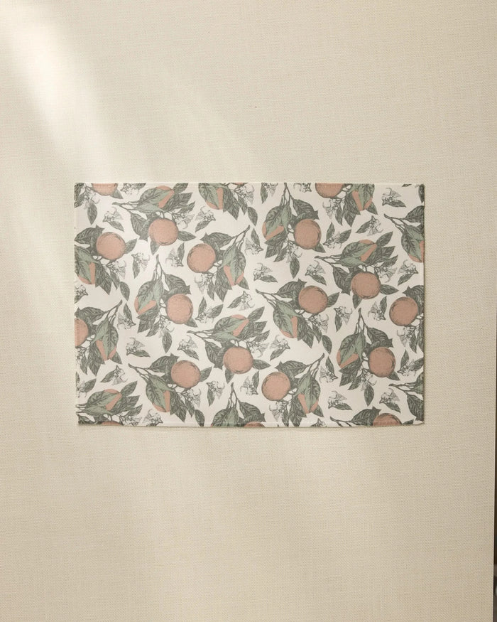 The Valencia rectangular sheet by Geometry, featuring an orange and green leaf pattern like recycled placemats, rests on a beige textured surface bathed in soft natural light from the left.
