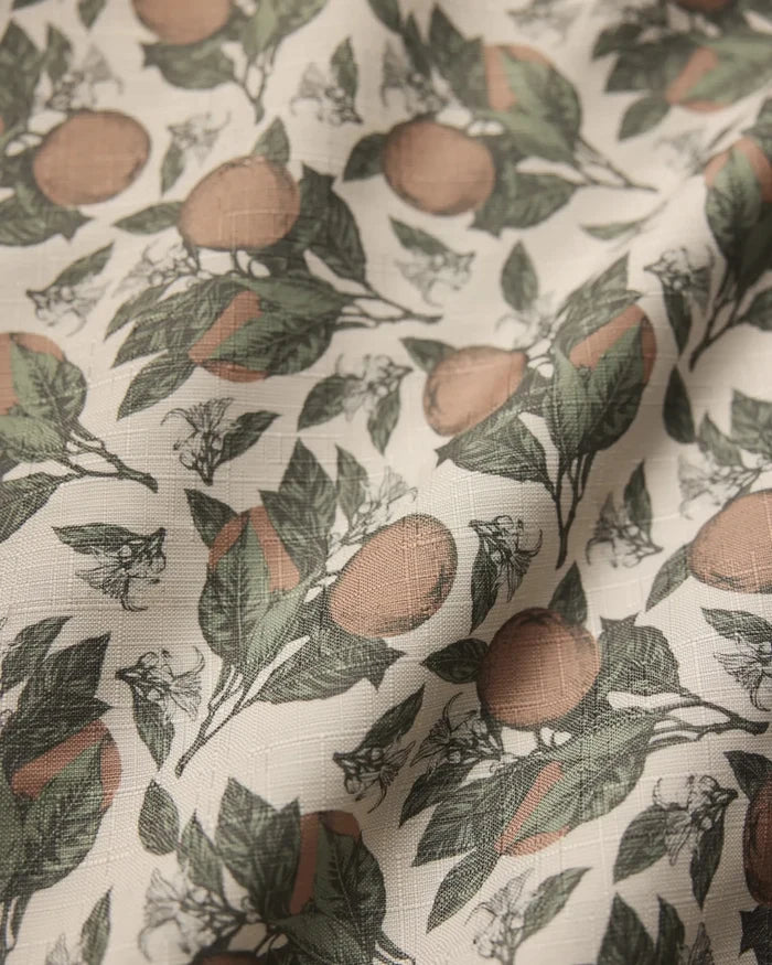 A close-up of Geometrys Valencia tablecloth highlights its lightweight, slightly wrinkled texture and botanical design with green leaves, brownish-orange fruits, and white flowers on a light background.