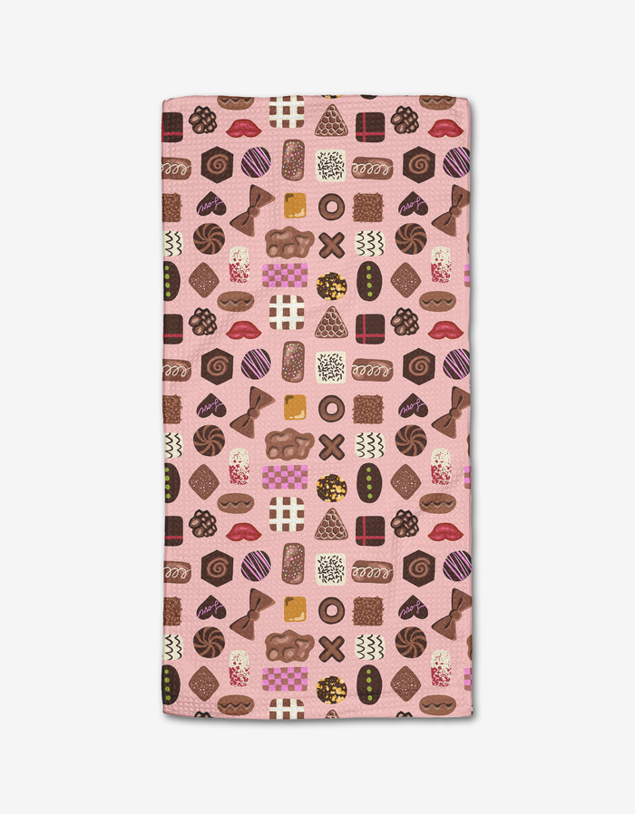 The Elisabeth Wing “Valentine’s Chocolates” Bar Towel features a pink background with a colorful pattern of assorted chocolates and candies. Made from Geoweave fabric, the rectangular towel is shown flat on a white surface.