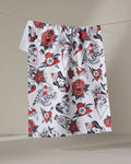 A white tea towel, “Valentine's Flash” by Cole Strem, hangs on a line. Made from recycled materials, its bold red, black, and gray tattoo-inspired design features roses, hearts, daggers, faces, and vintage “LOVE” banners.