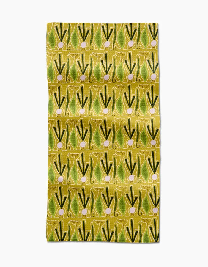 The Lizzie Clark Veg on Parade bar towel features a repeating pattern of green and yellow stylized leeks or onions with pinkish bulbs on a yellow background. Made from slightly wrinkled Geoweave fabric crafted from recycled materials.