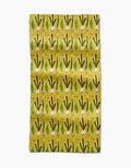 The Lizzie Clark Veg on Parade bar towel features a repeating pattern of green and yellow stylized leeks or onions with pinkish bulbs on a yellow background. Made from slightly wrinkled Geoweave fabric crafted from recycled materials.