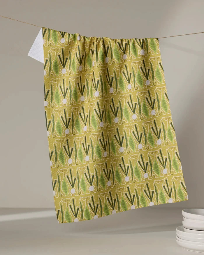 The "Veg on Parade" tea towel by Lizzie Clark, featuring a yellow background and green onion pattern, hangs on a line above a white surface with stacked white dishes in the corner.