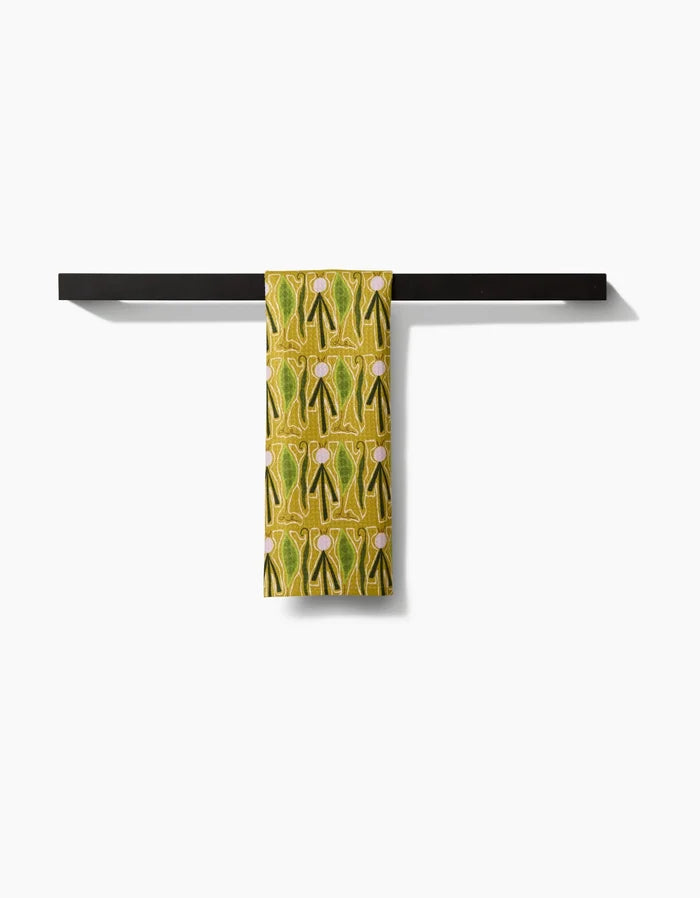 The Veg on Parade bar towel by Lizzie Clark, featuring a yellow-green pattern and made from recycled materials, is neatly draped over a minimalist black bar with a plain white backdrop.