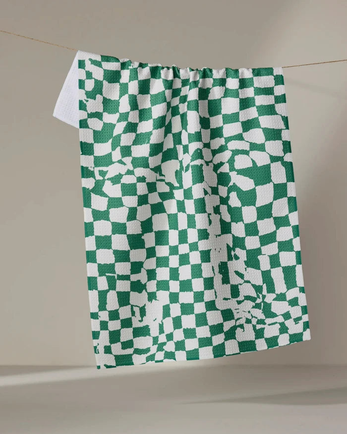 The Verdant kitchen towel by Geometry, featuring a green and white checkered pattern with scattered, abstract squares, hangs on a clothesline against a plain, neutral indoor background.